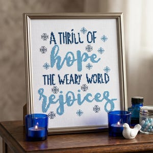 May include: A framed cross-stitch artwork with the words "A thrill of hope, the weary world rejoices" in blue script. The frame is silver. Two blue votive candles and a small white bird figurine sit on a wooden table.