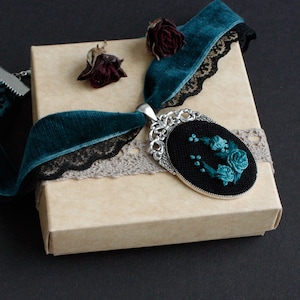 May include: A teal blue velvet ribbon with black lace trim and a silver pendant with a black and teal embroidered floral design. The pendant is in a gift box.