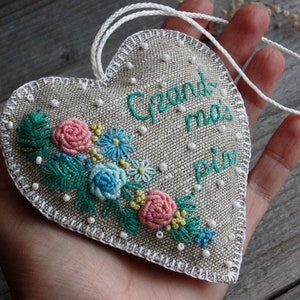 May include: A heart-shaped linen cushion with white stitching and a floral embroidery design. The cushion has a white cord attached and the text "Grand ma's pin" is embroidered on the front.