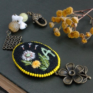 May include: A black felt brooch with embroidered flowers in white, yellow, and green, framed by yellow beads. It has a bronze-coloured floral charm and a decorative metal piece, plus a beaded chain and clasp. The brooch is oval-shaped.