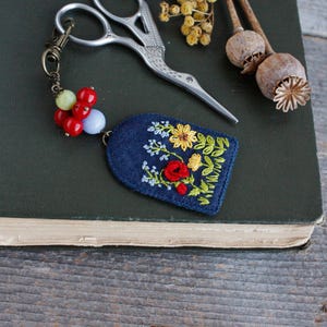 May include: A blue felt keychain with a floral embroidery design. The keychain features red, yellow, and blue flowers and green leaves. The keychain is attached to a silver-toned metal chain with a lobster clasp and a cluster of red, green, and blue beads.