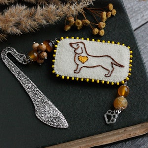 May include: A handmade bookmark featuring a dachshund design. The rectangular beige felt piece has a brown embroidered dog with a yellow heart. The bookmark has a silver metal end with an ornate design and a beaded charm with a paw print charm.