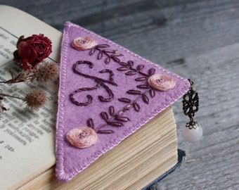 May include: A lavender felt bookmark with embroidered floral and initial details. The bookmark features a decorative floral design and a dangling charm with a pink gemstone. The bookmark is placed in an open book.