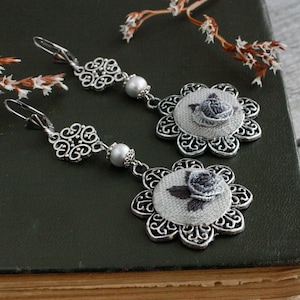 May include: A pair of silver-toned dangle earrings featuring a filigree flower frame, a single white pearl bead, and a central fabric inlay embroidered with a small grey rose. The earrings are equipped with lever-back closures for secure wear.