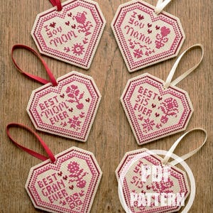 Könnte beinhalten: Sechs herzförmige Kreuzstich-Ornamente in Rosa und Creme. Jedes Ornament trägt eine andere Botschaft, darunter "I Love Mom", "Best Mom Ever" und "Best Grandma Ever". Sie haben Bänder zum Aufhängen.