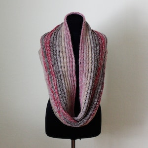 以下が含まれることがあります： ピンク、赤、グレー、ベージュの色合いのニットインフィニティスカーフ。黒いマネキンに展示されており、リブ編みの質感と色のバリエーションを示しています。柔らかく暖かいアクセサリーは、肌寒い季節に最適です。