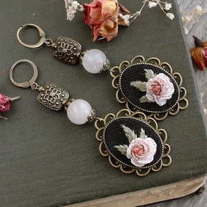 May include: A pair of earrings with a delicate rose embroidery on a black background, framed in an ornate gold-toned metal setting. Each earring features a white quartz bead and a decorative metal charm.