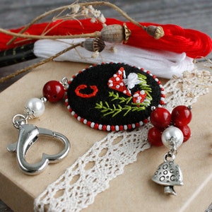 May include: A black felt keychain with a red and white embroidered mushroom design. The keychain has a silver heart-shaped clasp and a silver bell charm with a red bead and white pearl accents.