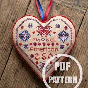 May include: Heart-shaped cross-stitch ornament with red, white, and blue embroidery. The design includes floral patterns and the text "My heart is all American." A red and blue ribbon is attached for hanging. The words "PDF PATTERN" are in a white circle.