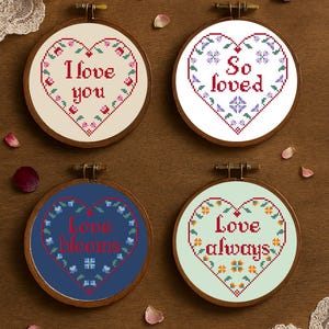 Op de afbeelding: Vier borduurringen met kruissteek met hartvormige ontwerpen. Elke ring heeft een andere boodschap: "I love you", "So loved", "Love blooms" en "Love always". De ontwerpen zijn in rood, blauw en groen, met bloemaccenten.