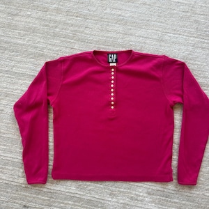 Könnte beinhalten: Ein langärmeliges, fuchsiafarbenes Pullover-Shirt von GAP mit rundem Halsausschnitt und einer vertikalen Knopfleiste in der vorderen Mitte. Die Leiste ist mit einer Reihe von acht kleinen, hellen Knöpfen verziert. Das Kleidungsstück hat einen geraden Saum.