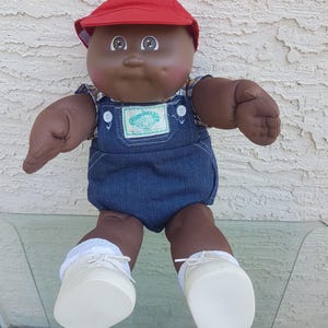 Peut inclure: Poupée Cabbage Patch Kids à la peau foncée, portant une casquette rouge, une salopette en jean et des chaussures blanches. La poupée a une expression souriante et est assise sur une surface en verre. Le logo Cabbage Patch Kids est visible.
