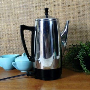 Op de afbeelding: Een vintage chromen koffiepercolator met een zwarte handgreep en basis. De koffiepot heeft een tuit en een zwarte bovenkant. Twee lichtblauwe keramische kopjes en een kleine plant staan op de achtergrond.