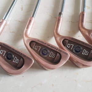 以下が含まれることがあります： ローズゴールド色のヘッドとシルバーのシャフトが付いた4本のゴルフクラブのセット。クラブヘッドには「50」と「SOLO-SEA II SUPER MO. OF INERTIA」の文字が書かれています。シャフトには「BERYLLIUM」の文字が印刷されています。