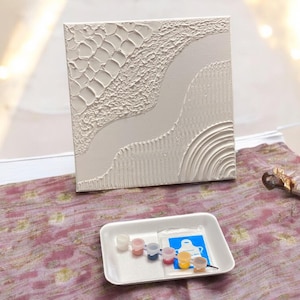 May include: A square white canvas featuring raised textured patterns including a grid, a smooth winding path, and concentric semi-circles. Below the canvas sits a white tray containing six small paint pots and a blue packet with a white apron graphic.