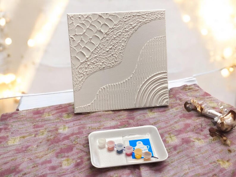 May include: A square white canvas featuring raised textured patterns including a grid, a smooth winding path, and concentric semi-circles. Below the canvas sits a white tray containing six small paint pots and a blue packet with a white apron graphic.