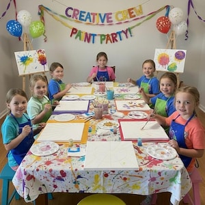 May include: Six children wearing colorful aprons sit around a table covered in a paint-splattered cloth, each with a blank canvas, paintbrushes, and a palette. A banner above reads CREATIVE CREW PAINT PARTY, with balloons on wooden easels nearby.