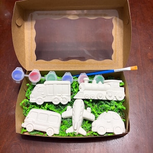 May include: A craft kit featuring a cardboard box with a clear window, containing white plaster vehicle figurines: a van, a train, a car, a bus, and an airplane. The kit includes small paint pots in red, green, blue, and purple, along with a paintbrush.