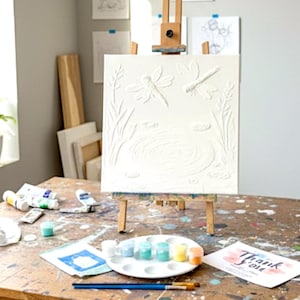 May include: A square white canvas with a raised relief design of two dragonflies hovering over a pond with reeds, mounted on a wooden easel. The canvas features a textured surface with monochromatic details depicting a nature scene with insects.