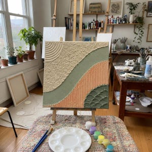 May include: A square canvas painting featuring textured sections in cream, sage green, and peach. The design includes a circular ripple pattern in the top left, a smooth diagonal sage stripe, vertical lines in peach, and a scalloped green texture.