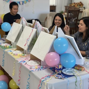May include: A table set for a painting party featuring three white canvases on wooden easels, each displaying a raised hot air balloon design. The table is decorated with pink, blue, and yellow balloons, white paint palettes, and colorful ribbons.