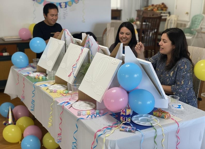 May include: A table set for a painting party featuring three white canvases on wooden easels, each displaying a raised hot air balloon design. The table is decorated with pink, blue, and yellow balloons, white paint palettes, and colorful ribbons.