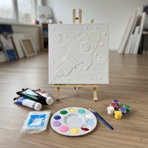 May include: A white canvas with a raised rocket ship design on a small wooden easel. Paint tubes, a palette with various colors, a paintbrush, and small paint pots are arranged nearby, suggesting a DIY painting kit.