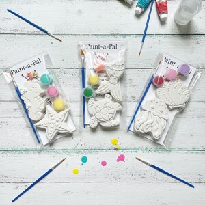 May include: Three Paint-a-Pal craft kits in clear plastic packaging. Each kit contains white plaster sea-themed figures, a small paintbrush, and three small paint pots. The figures include a seahorse, starfish, mermaid tail, turtle, jellyfish, and seashell.