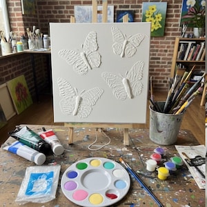 May include: A square white canvas on a wooden easel featuring four raised, textured butterfly sculptures in white. The canvas is displayed on a wooden table surrounded by various art supplies, including paint tubes, a round plastic palette with colorful paints, and a metal container filled with assorted paintbrushes.