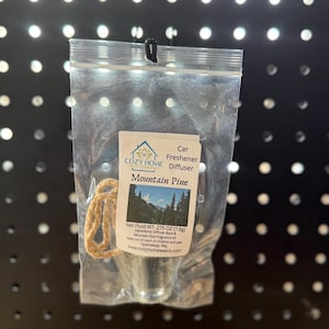 May include: A clear plastic bag containing a car air freshener. The label reads "Mountain Pine" with a mountain scene. The net fluid weight is 0.275 oz (7.8g). A beige string is attached for hanging.