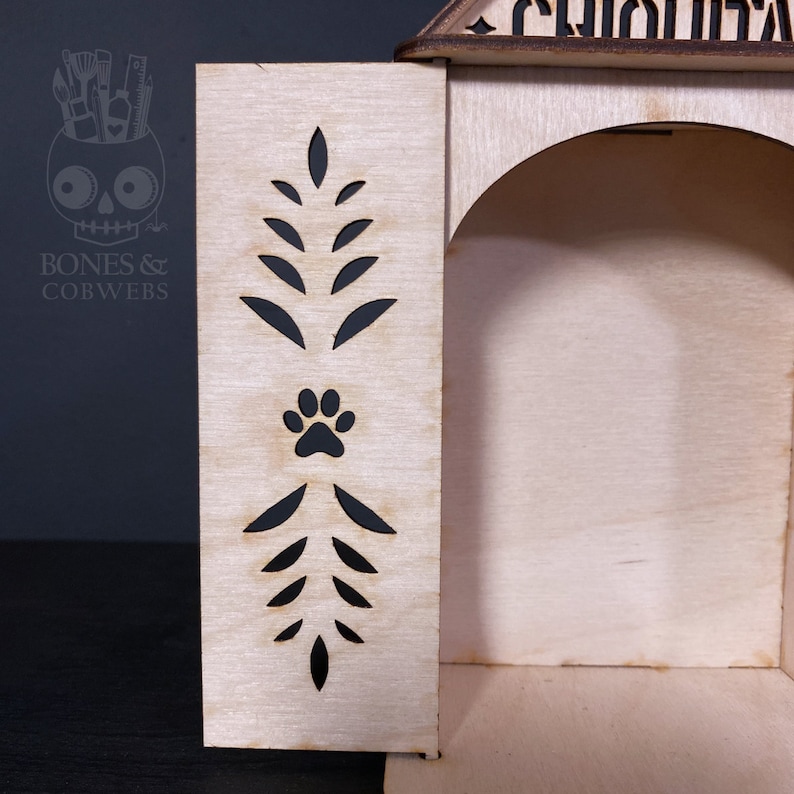 May include: A light-coloured wooden structure featuring a laser-cut side panel with a symmetrical leaf pattern and a central paw print cutout. The structure has an arched opening and a roof section with partially visible black lettering.