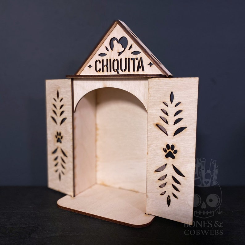 May include: A light-coloured wooden shrine with an arched opening and two hinged doors. The triangular top features a cutout heart with a silhouette of a dog and the name CHIQUITA. Each door has laser-cut leaf patterns and a paw print design.