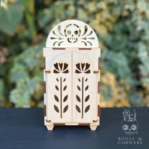 May include: A wooden box with a skull and floral design. The box has two doors that open. The box is made of light wood and has a brown stain on the edges.