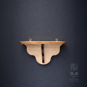 May include: A wooden wall shelf with a scalloped bottom edge and a dark brown support. The shelf is made of light-colored wood with a natural finish. Two small gold-colored rings are attached to the top edge for hanging. The shelf is against a dark blue background.