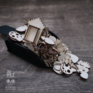 May include: A black scoop overflowing with a variety of wooden cutouts, including hearts, flowers, skulls, and a small wooden frame. The cutouts are in various sizes and shapes, and they are all made of unfinished wood.