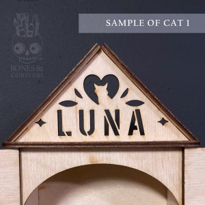 May include: A triangular wooden sign featuring the laser-cut name LUNA in bold capital letters. Above the name is a cutout of a cat silhouette inside a heart shape, flanked by small leaf motifs and four-pointed stars on the edges of the frame.