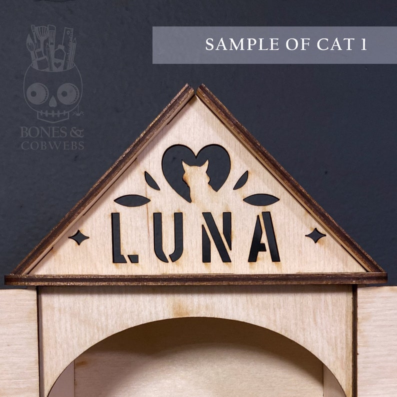 May include: A triangular wooden sign featuring the laser-cut name LUNA in bold capital letters. Above the name is a cutout of a cat silhouette inside a heart shape, flanked by small leaf motifs and four-pointed stars on the edges of the frame.