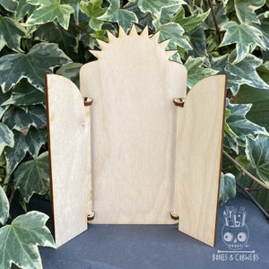 May include: A wooden three-panel display stand with a sunburst design at the top. The stand is made of unfinished wood and has a natural color.
