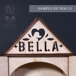 May include: A triangular wooden sign featuring a laser-cut silhouette of a dog inside a heart shape, with the name BELLA cut out in block letters. The sign has dark stained edges and a light wood finish, with small star accents on each side.
