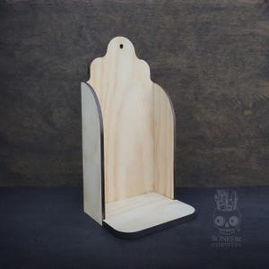 May include: A wooden wall shelf with a decorative top and a small shelf at the bottom. The shelf is made of light-colored wood with dark brown edges. The shelf has a hole at the top for hanging. The shelf is approximately 10 inches tall.