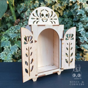 May include: A wooden Day of the Dead altar with a skull design on the top and floral cutouts on the doors. The doors are open, revealing an empty interior.