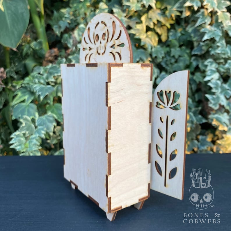 May include: A wooden box with a skull design on the top and a floral design on the side. The box is made of light wood and has a natural finish. The box is closed and the lid is slightly ajar. The box is sitting on a black surface. "Bones & Cobwebs" is printed on the bottom of the image.