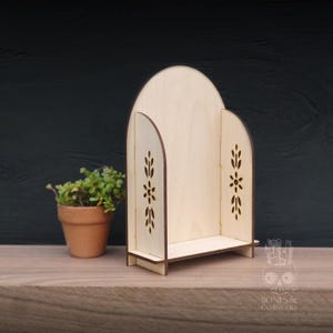 Puede incluir: Una estantería de madera clara con una parte superior arqueada y recortes florales en los lados. La estantería tiene una base pequeña y una zona de estantería. Una pequeña planta en maceta está a la izquierda.