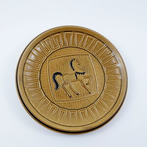 Può includere: Un piatto in ceramica marrone con un disegno di cavallo bianco e nero al centro. Il piatto ha un bordo decorativo di quadrati ripetuti.