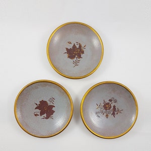 Könnte beinhalten: Drei kleine, runde, dekorative Teller mit goldenen Rändern und Blumendesigns. Die Teller sind hellgrau mit braunen Blumendesigns.
