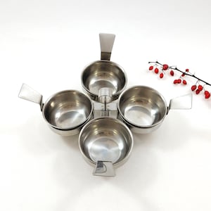 Peut inclure: Un plat de service en acier inoxydable avec quatre bols ronds, chacun avec une poignée rectangulaire. Les bols sont disposés en forme de fleur. Une branche de baies rouges est en arrière-plan.