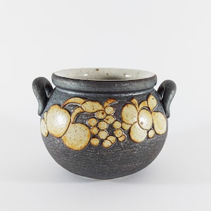 Könnte beinhalten: Ein kleiner, dunkelgrauer Keramiktopf mit zwei Henkeln. Der Topf hat eine weiße Glasur mit einem Design aus Trauben und Blumen.