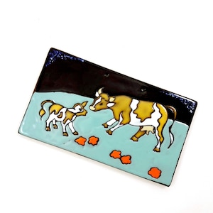 Op de afbeelding: Een blauwe en bruine keramische tegel met een geschilderd beeld van een koe en een kalf. De koe is bruin en wit met een zwarte neus en hoeven. Het kalf is wit met bruine vlekken. Er zijn oranje bloemen geschilderd op de tegel.