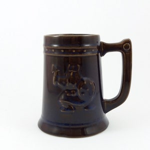 May include: A brown ceramic beer stein featuring a raised design of a Viking holding an axe. The stein has a brown handle and a brown rim.