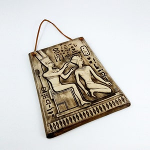 K&ouml;nnte beinhalten: Eine braune Keramik-Wandplatte mit einem erhabenen Reliefdesign von zwei Figuren im alten &auml;gyptischen Stil. Die Figuren tragen traditionelle Kleidung und Kopfbedeckungen. Die Platte hat einen Seilhalter und ist mit Hieroglyphen verziert.